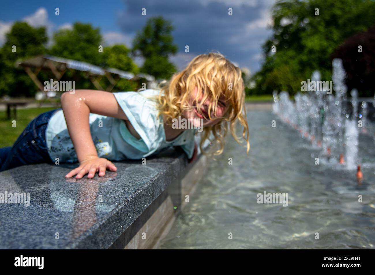 una ragazza dai capelli ricci immerge la mano nell'acqua fredda di una fontana. E' in jeans e una camicia azzurra, e il cielo blu e' limpido. Foto Stock