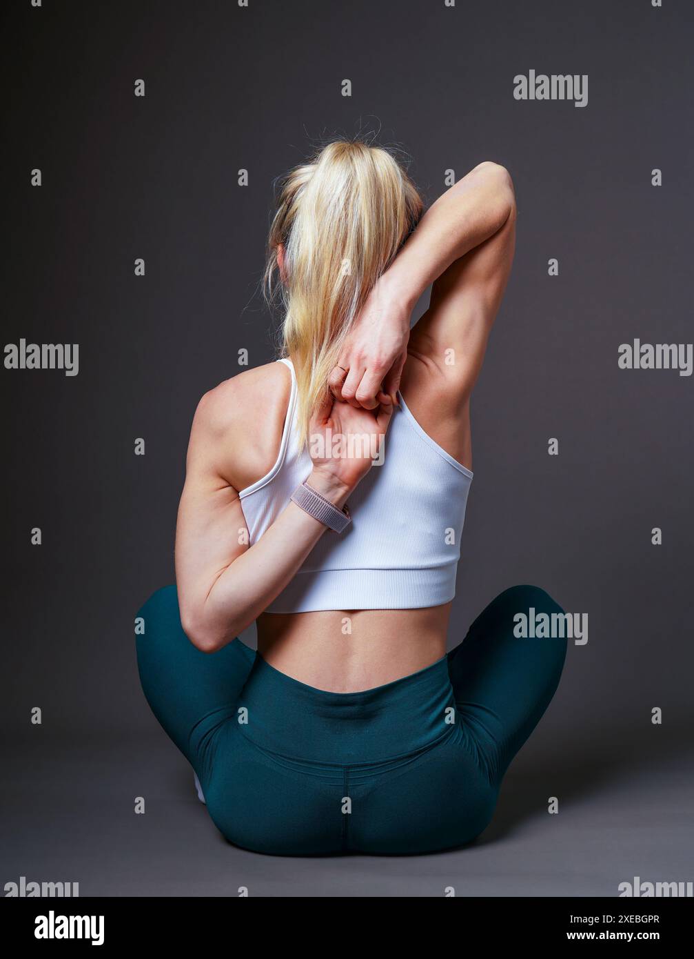 Una giovane ragazza europea con i capelli biondi in una maglietta bianca e pantaloni verdi sta facendo yoga. Mani nella serratura dietro la schiena. Su sfondo grigio. Atl Foto Stock