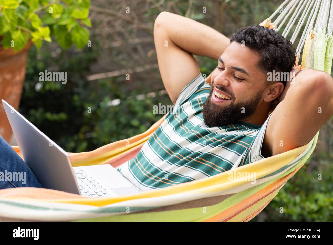 Utilizzando un computer portatile e sorridente, l'uomo si rilassa sull'amaca all'aperto Foto Stock