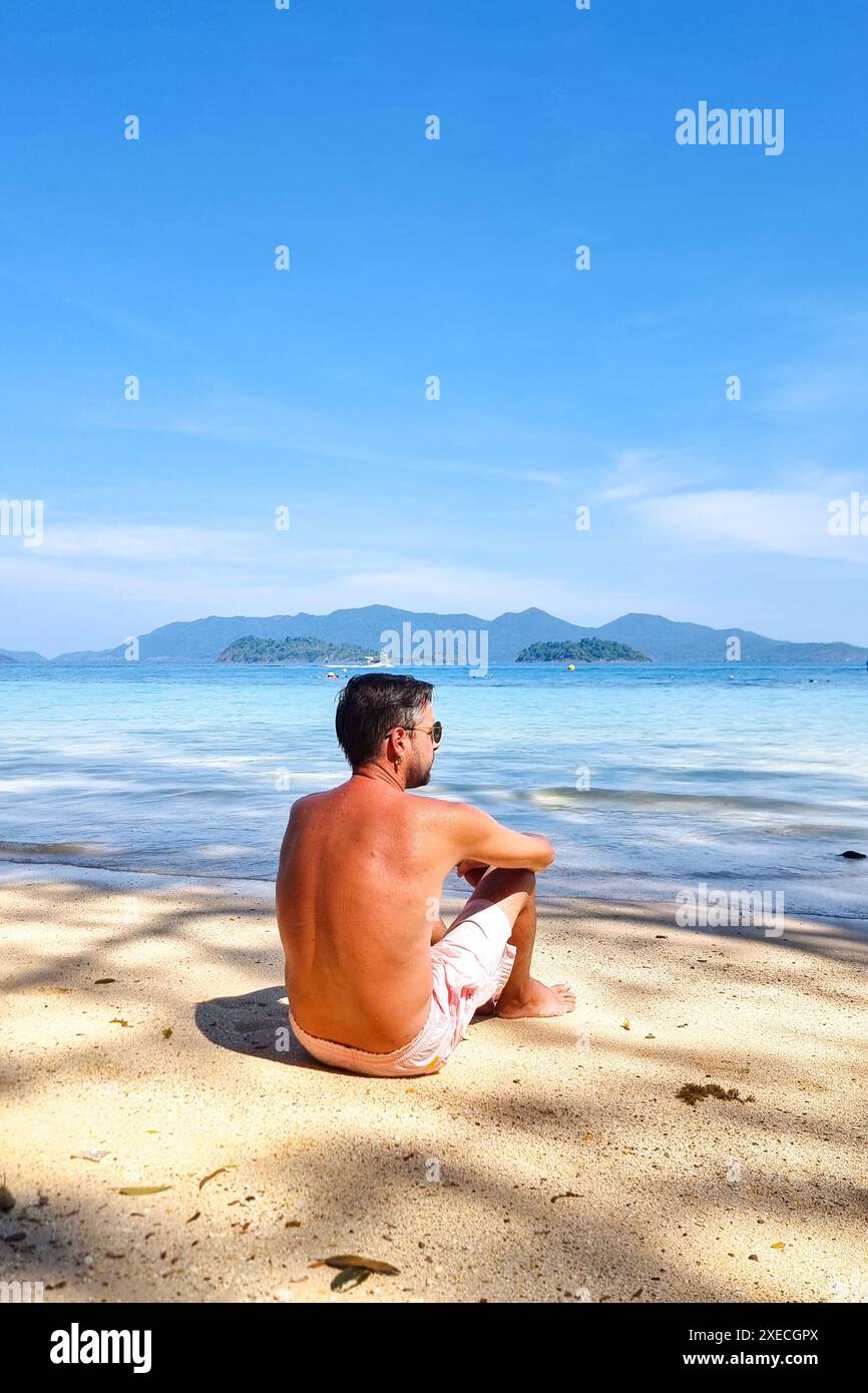 Un uomo con un abbigliamento casual si siede sulla spiaggia sabbiosa, il suo sguardo fisso sulla vasta distesa dell'oceano davanti a lui Foto Stock