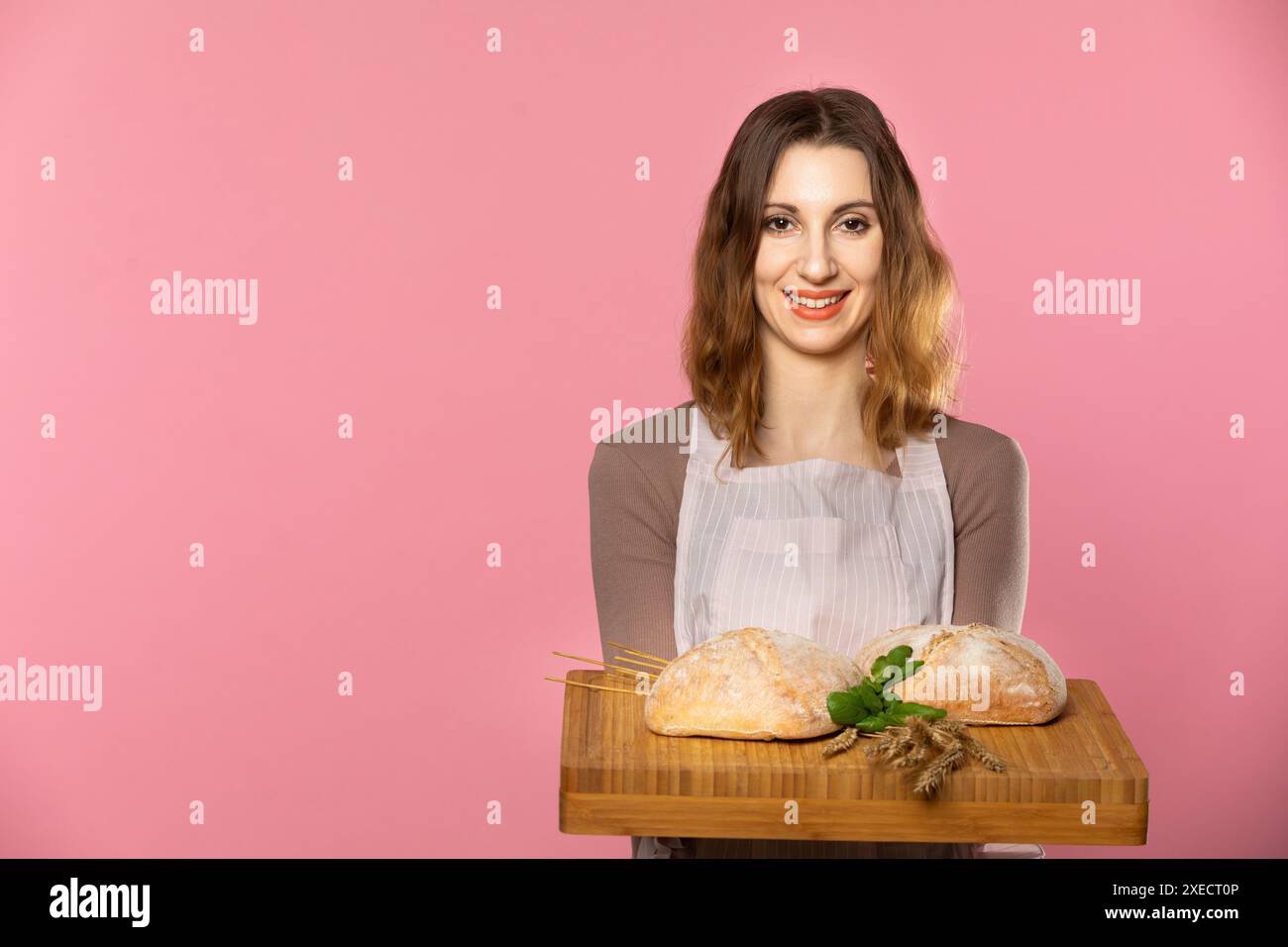 Una giovane e attraente venditrice tiene due soffici pani decorati con erbe e orecchie di grano maturo su una tavola di legno. Senza cuciture Foto Stock