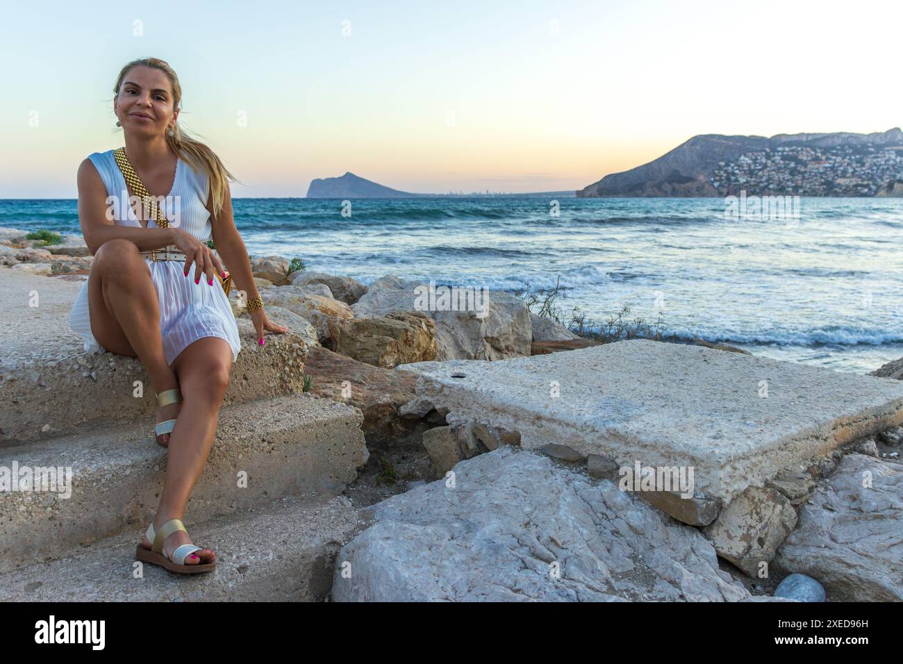 Questa immagine cattura una bella donna bionda che posa serenamente in riva al mare. È elegantemente vestita con un vestito bianco senza maniche Foto Stock