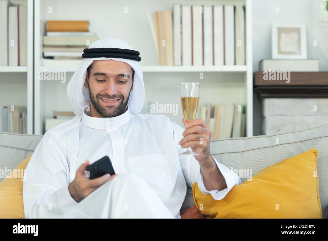 Ritratto di un uomo arabo sorridente che regge un bicchiere di champagne e usa il cellulare a casa Foto Stock