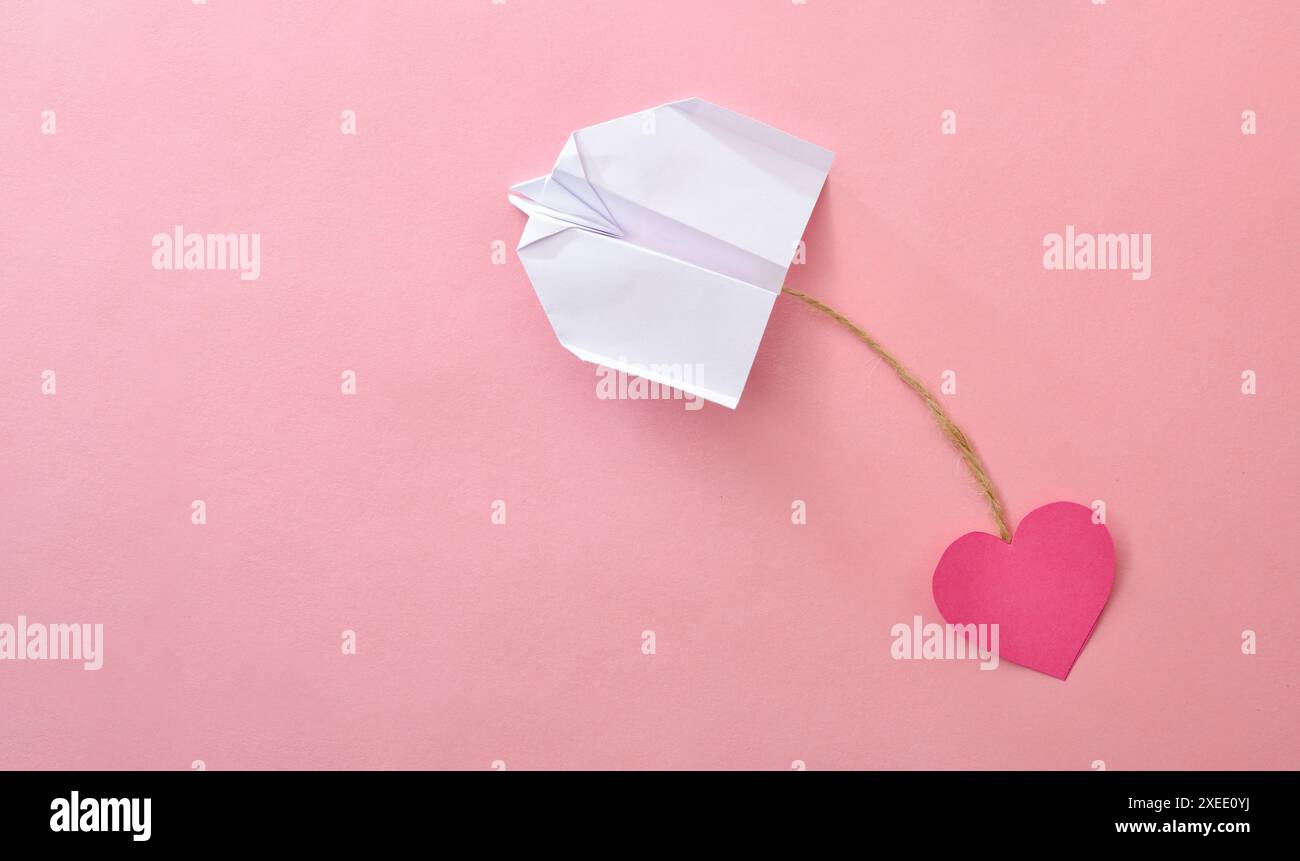 Concetto di amore per viaggiare in aereo con un aereo di carta che tira una fessura di carta a forma di cuore legata da una corda di iuta su sfondo rosa. Foto Stock