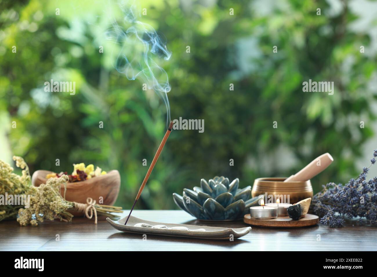 Bastone di incenso che si fonde nel supporto, candele accese, fiori secchi e ciotola tibetana per cantare su un tavolo di legno all'aperto Foto Stock