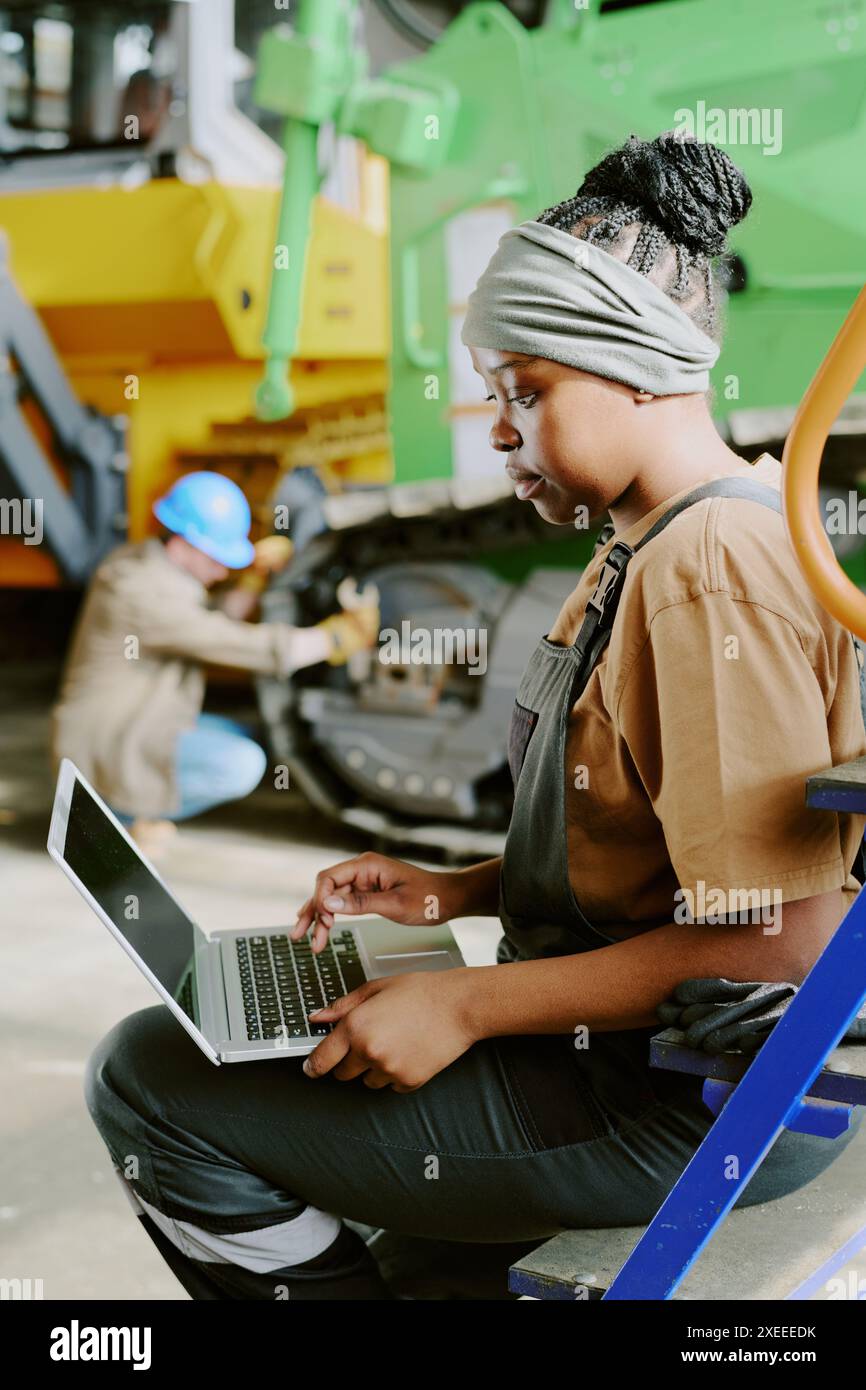 Immagine verticale di una giovane donna afroamericana che lavora in una fabbrica moderna controllando i dati su un computer portatile mentre il suo collega ripara la macchina in background Foto Stock