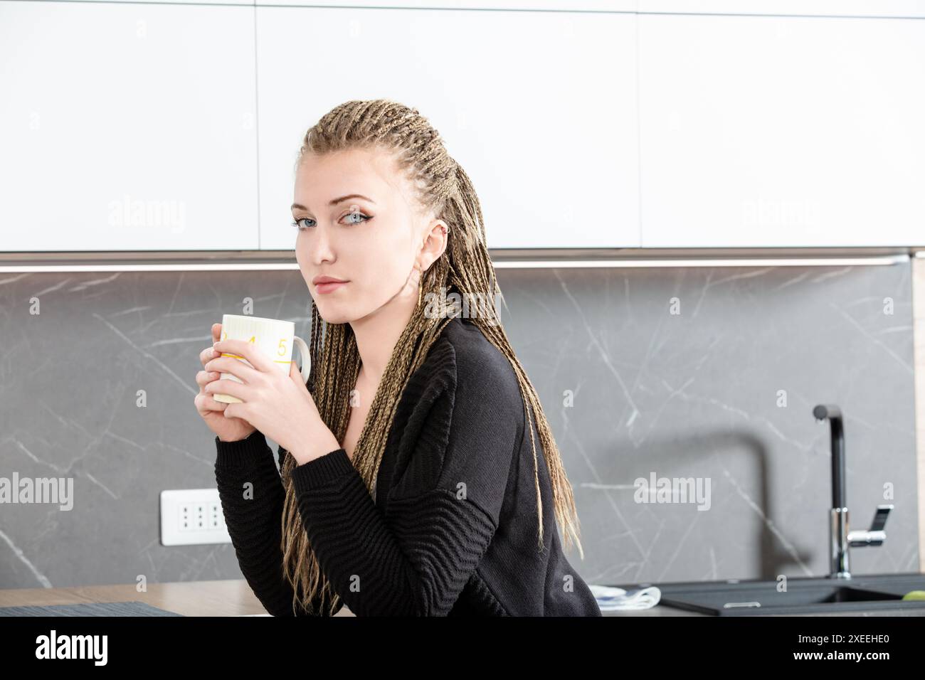 Una giovane donna con trecce bionde si gode una tranquilla pausa caffè nella sua moderna cucina, creando un'atmosfera accogliente e serena Foto Stock