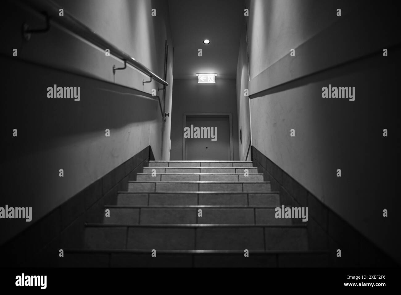 Gradini che precedono e un semaforo alla fine del corridoio con un cartello luminoso di uscita. Un corridoio con gradini, illuminati da lampade, ringhiere, porta all'en Foto Stock