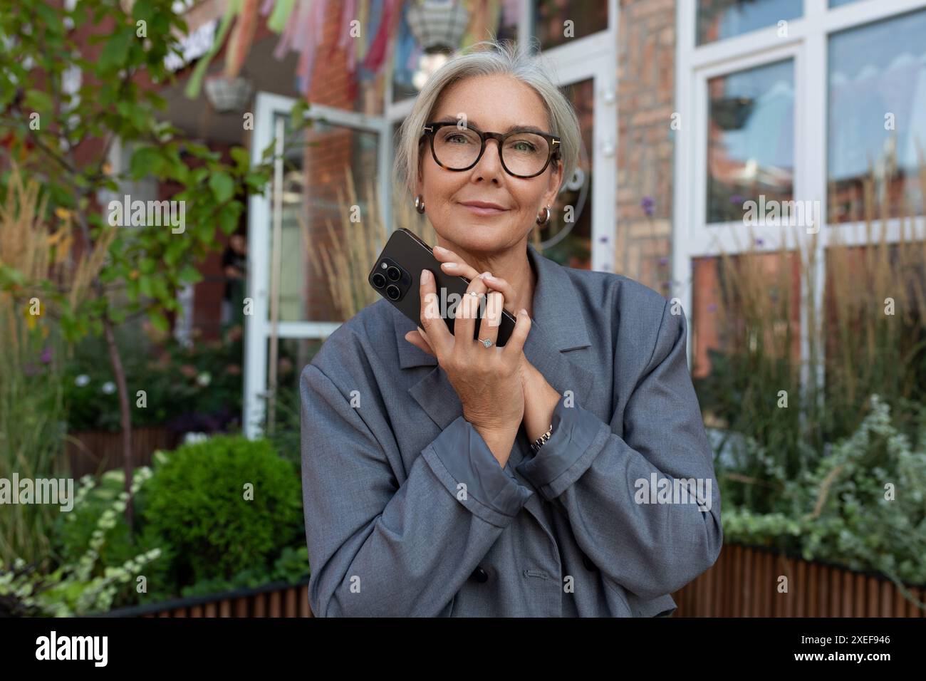 Una donna anziana ben curata con capelli e occhiali grigi è vestita con una giacca e un jeans grigi, tenendo in mano uno smartphone a. Foto Stock