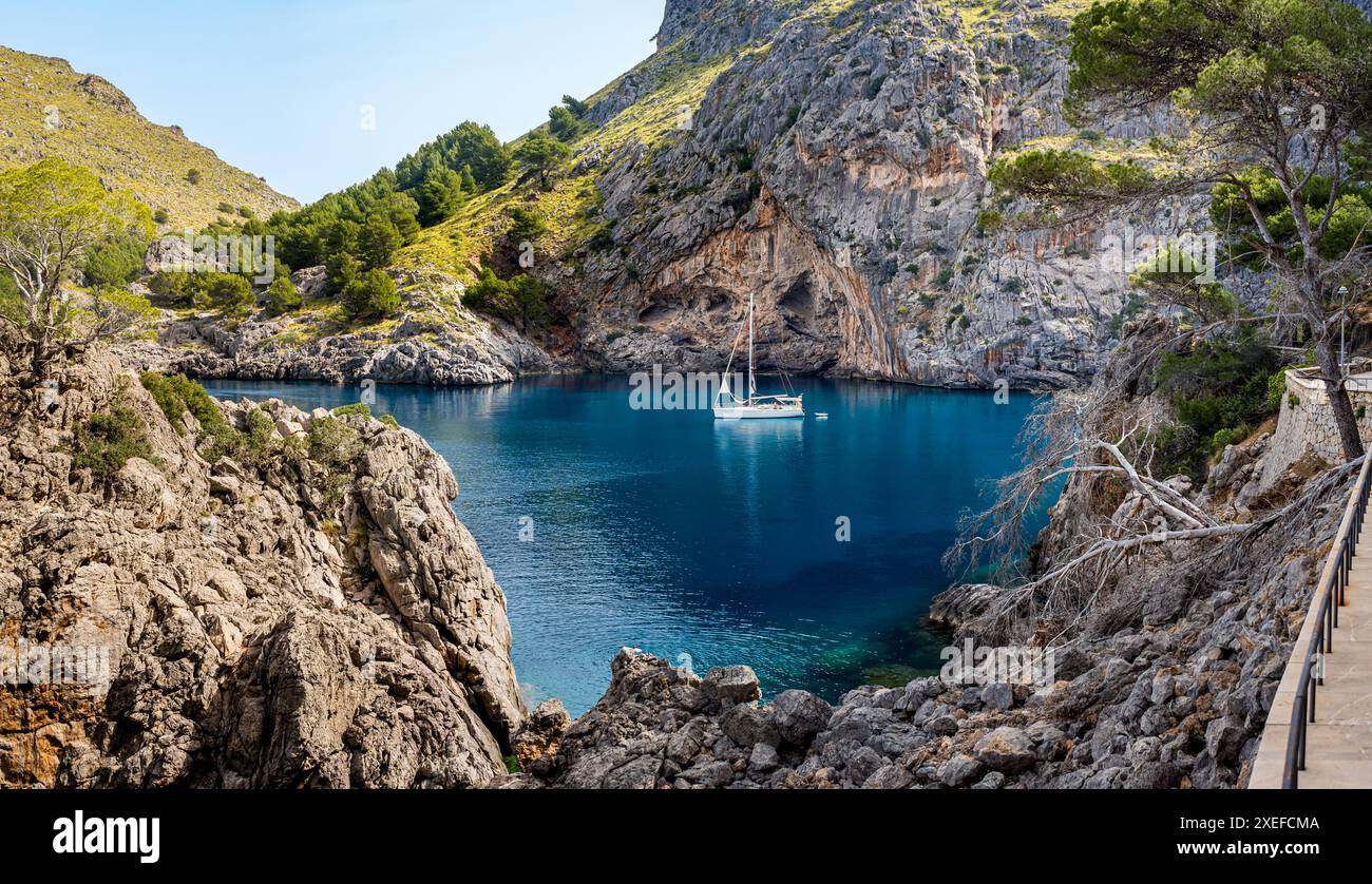 Il fascino tranquillo della baia di Cala de sa Calobra, che offre serene acque blu, una barca a vela ormeggiata e la montagna el Turmàs sullo sfondo, invitante a esplorare. Foto Stock