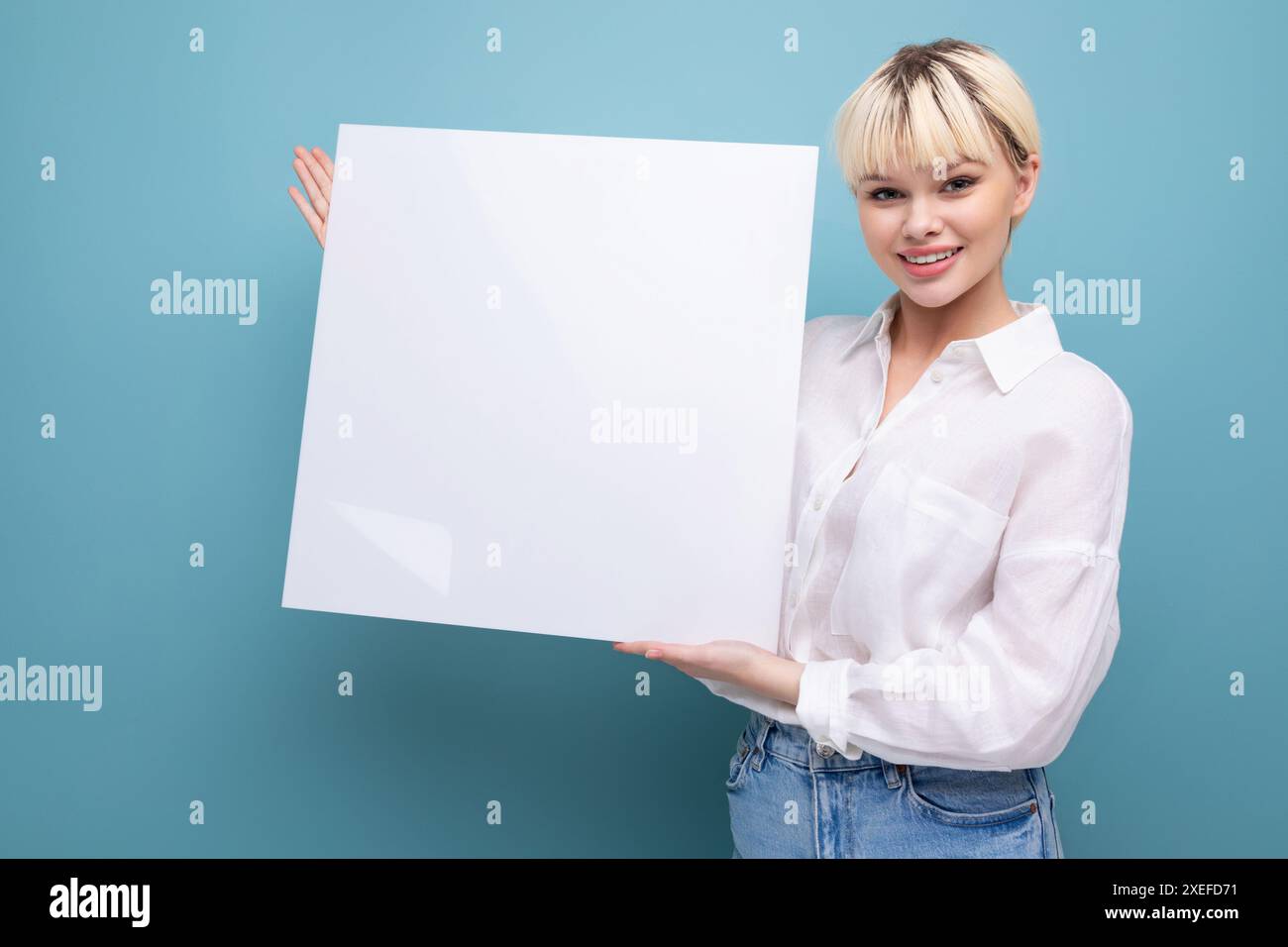 Giovane donna bionda di successo in ufficio con una camicia bianca e jeans tiene un pezzo di carta per inserire un annuncio pubblicitario. Foto Stock
