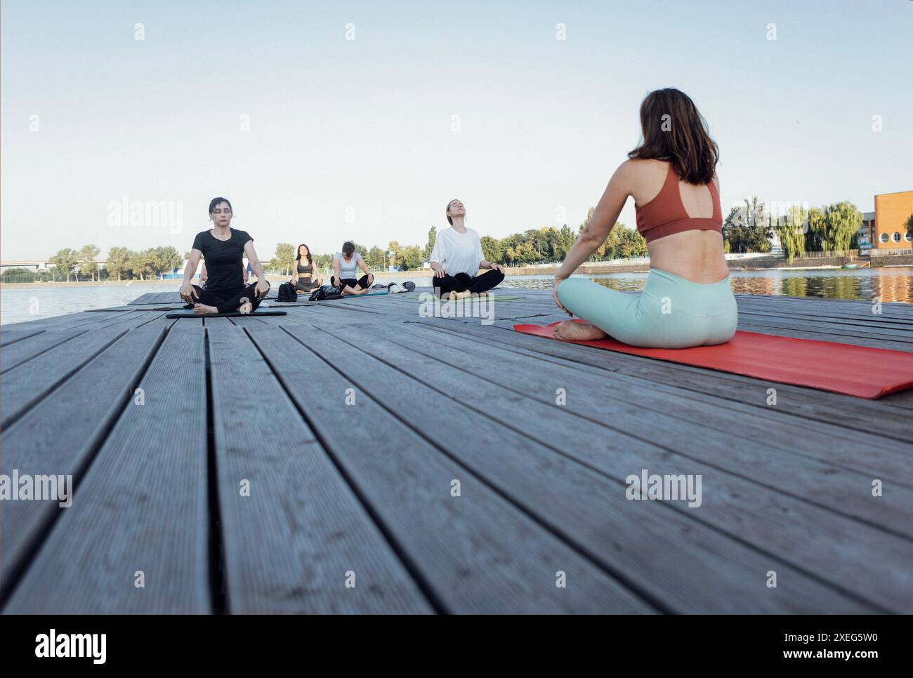 Lezione di yoga all'aperto con istruttore vicino al fiume. Le giovani donne siedono a gambe incrociate sui tappetini e meditano su un molo di legno. Foto Stock