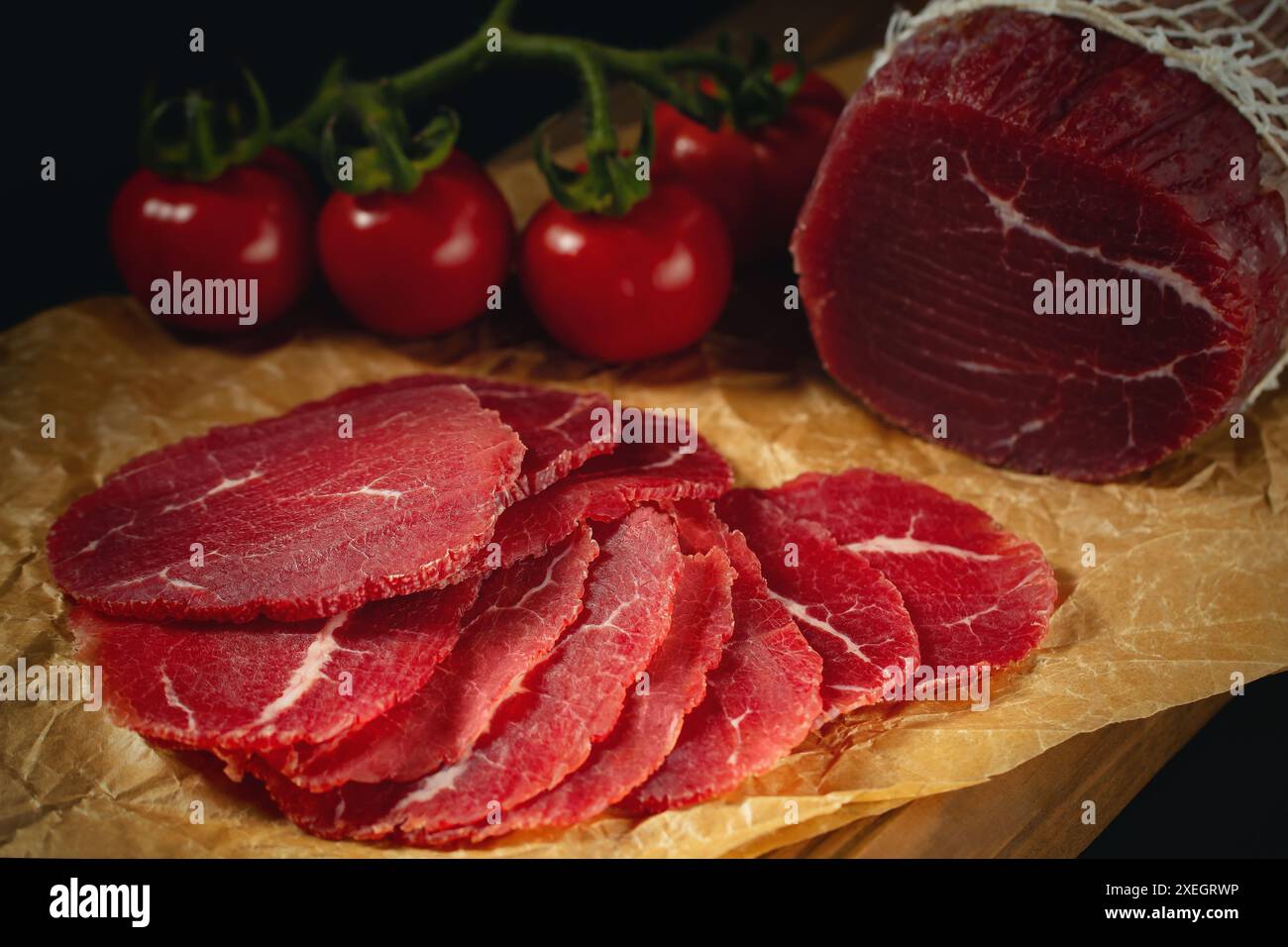 Bresaola intera e tagliata su carta su un tagliere con pomodori. Foto Stock
