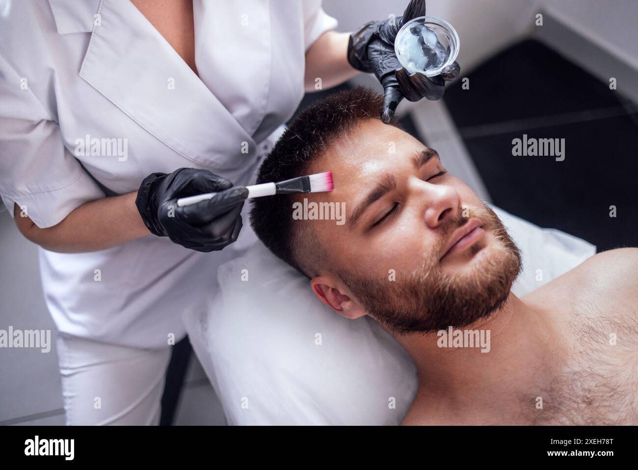 Il maestro di bellezza applica una maschera ringiovanente al viso maschile dei clienti. Sollevamento della pelle Foto Stock