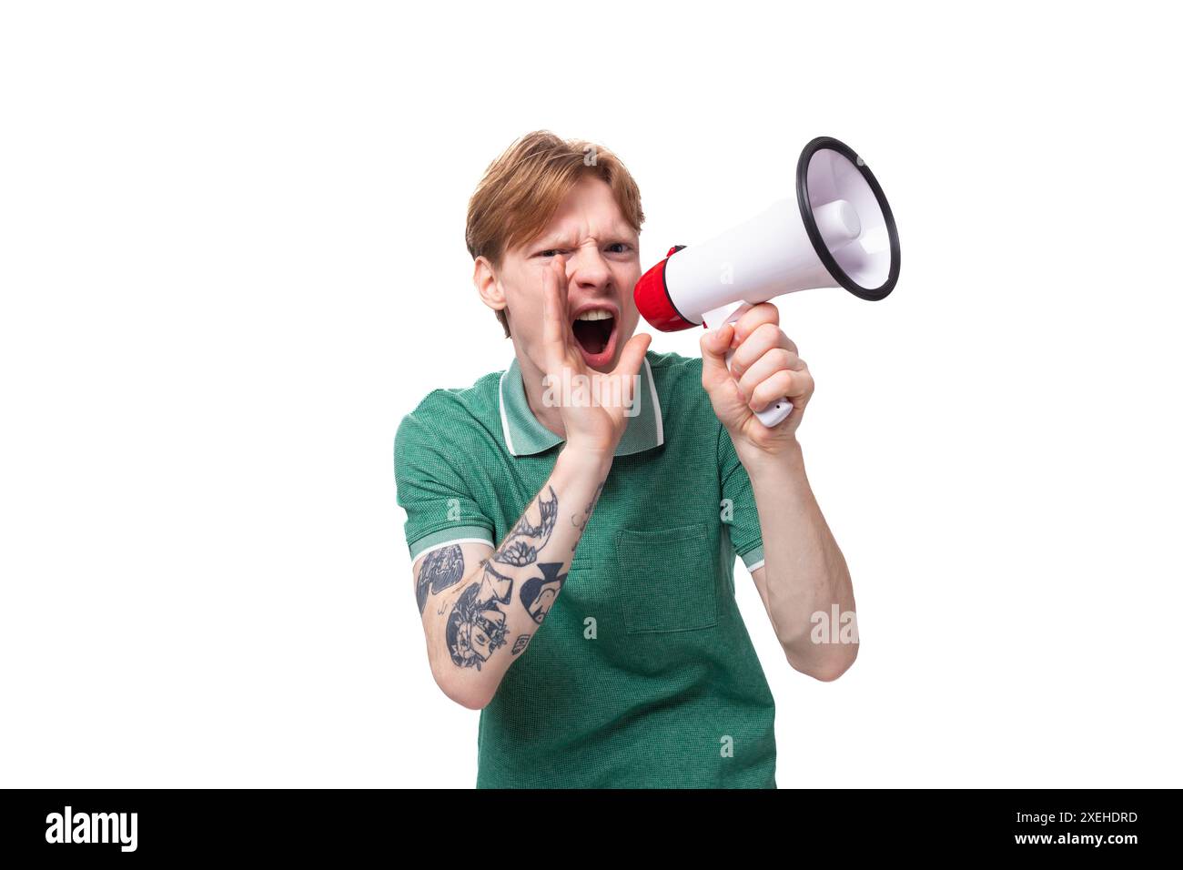 Il ritratto di un uomo dai capelli rossi sorpreso con una t-shirt verde con un tatuaggio sul braccio urla notizie in un altoparlante Foto Stock