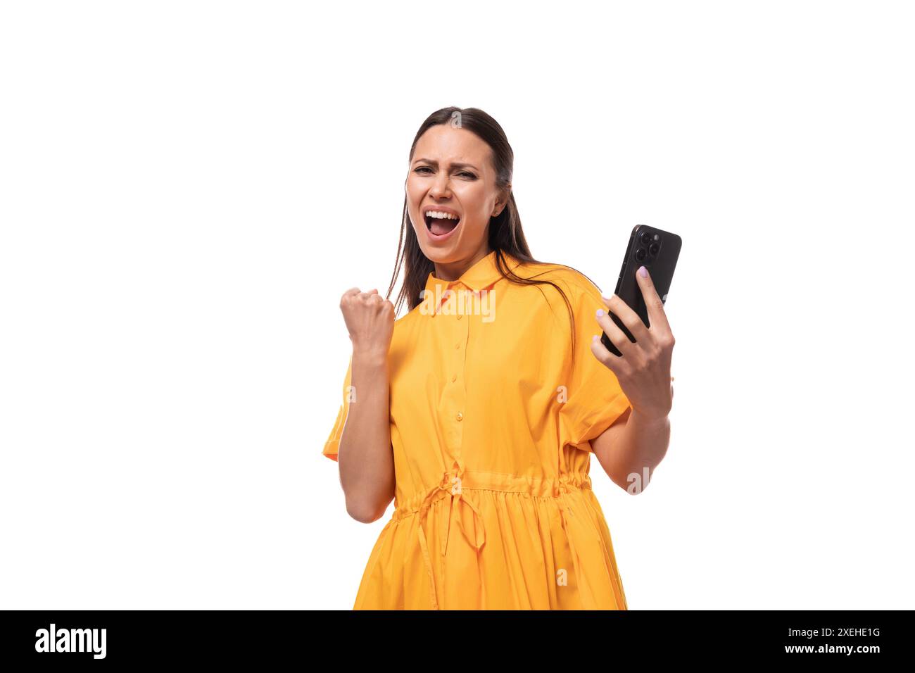 La giovane donna bruna caucasica con i capelli lunghi vestita con un vestito estivo giallo gioisce di successo tenendo in mano un telefono Foto Stock