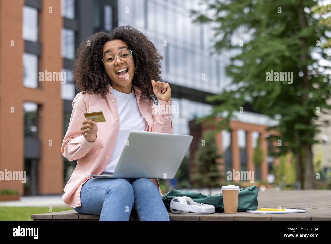 Giovane studentessa felice che tiene in mano la carta di credito mentre utilizza un laptop all'aperto vicino al campus universitario. Concetto di shopping online, istruzione e vita studentesca. Foto Stock