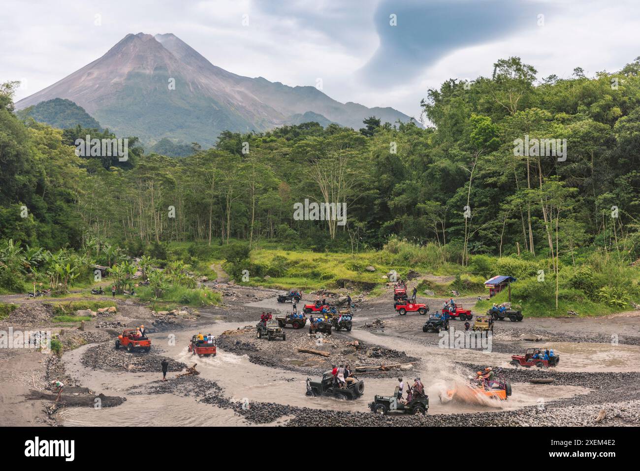 I veicoli commerciali sportivi corrono attraverso l'acqua e il fango con la vetta del Monte Merapi sullo sfondo nella parte centrale di Giava, Indonesia Foto Stock