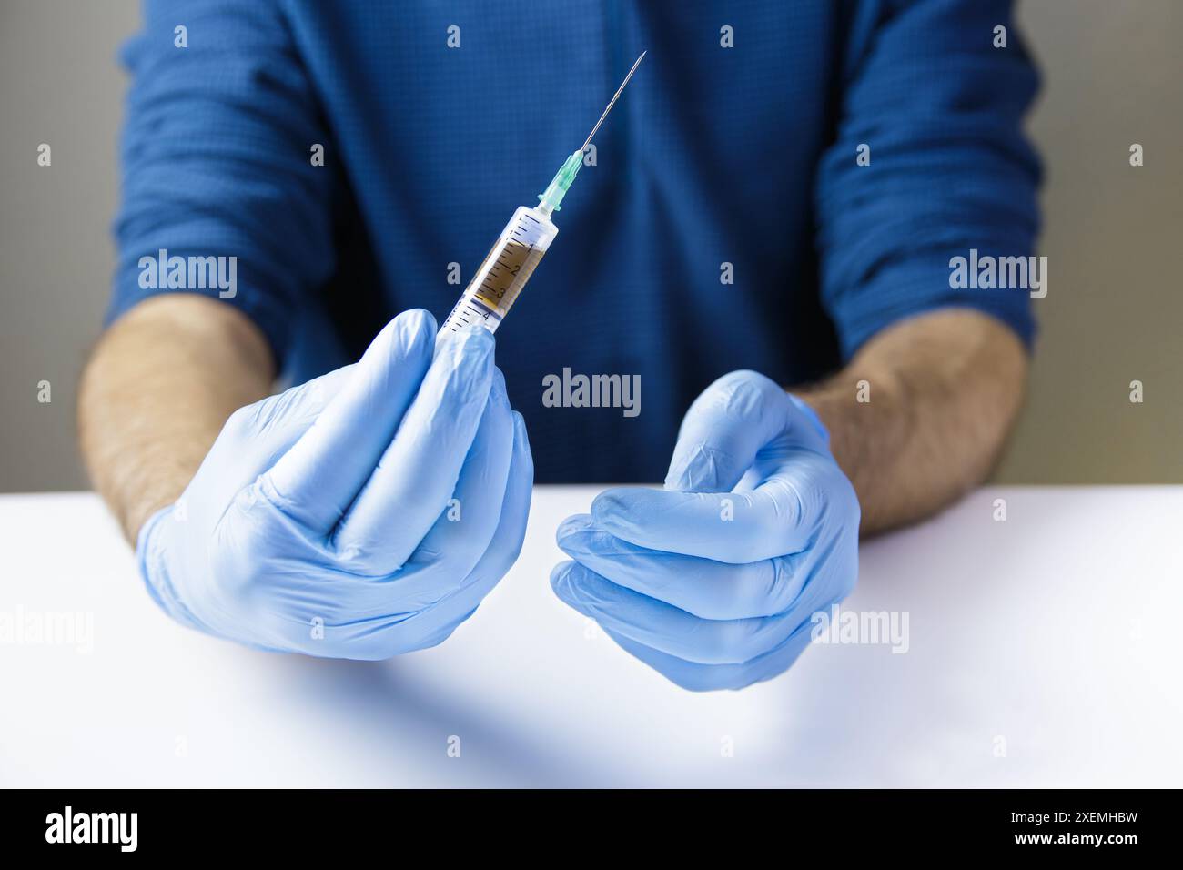 Le mani del medico o dell'infermiere, su un tavolo bianco e indossando guanti blu, tengono una siringa con un ago contenente un farmaco iniettabile all'interno. Orizzontale Foto Stock