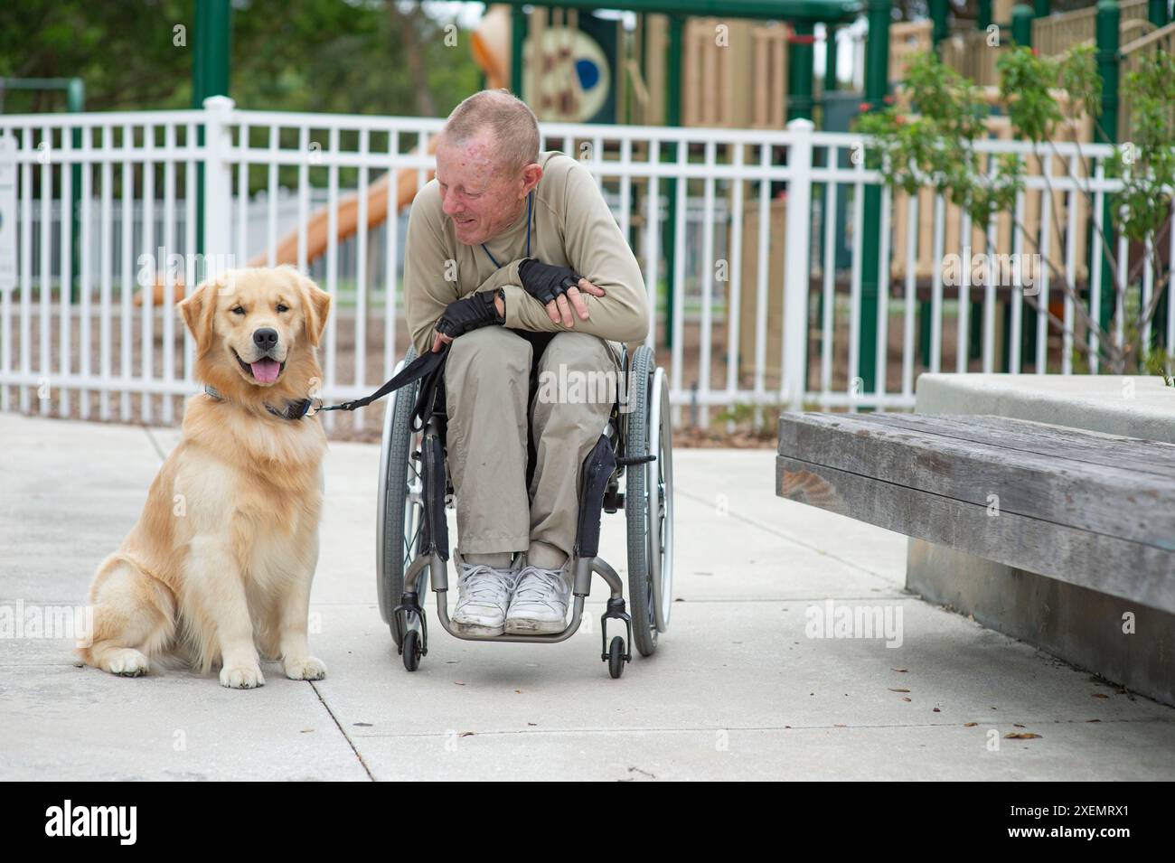 Uomo in sedia a rotelle con il suo cane di servizio in un parco giochi per bambini, un ritratto informale con l'uomo che guarda il cane Foto Stock