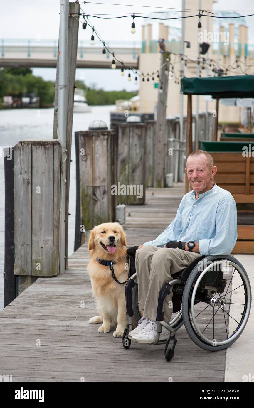 Ritratto informale all'aperto di un uomo in sedia a rotelle con il suo cane di servizio seduto su una passerella lungo un lungomare Foto Stock