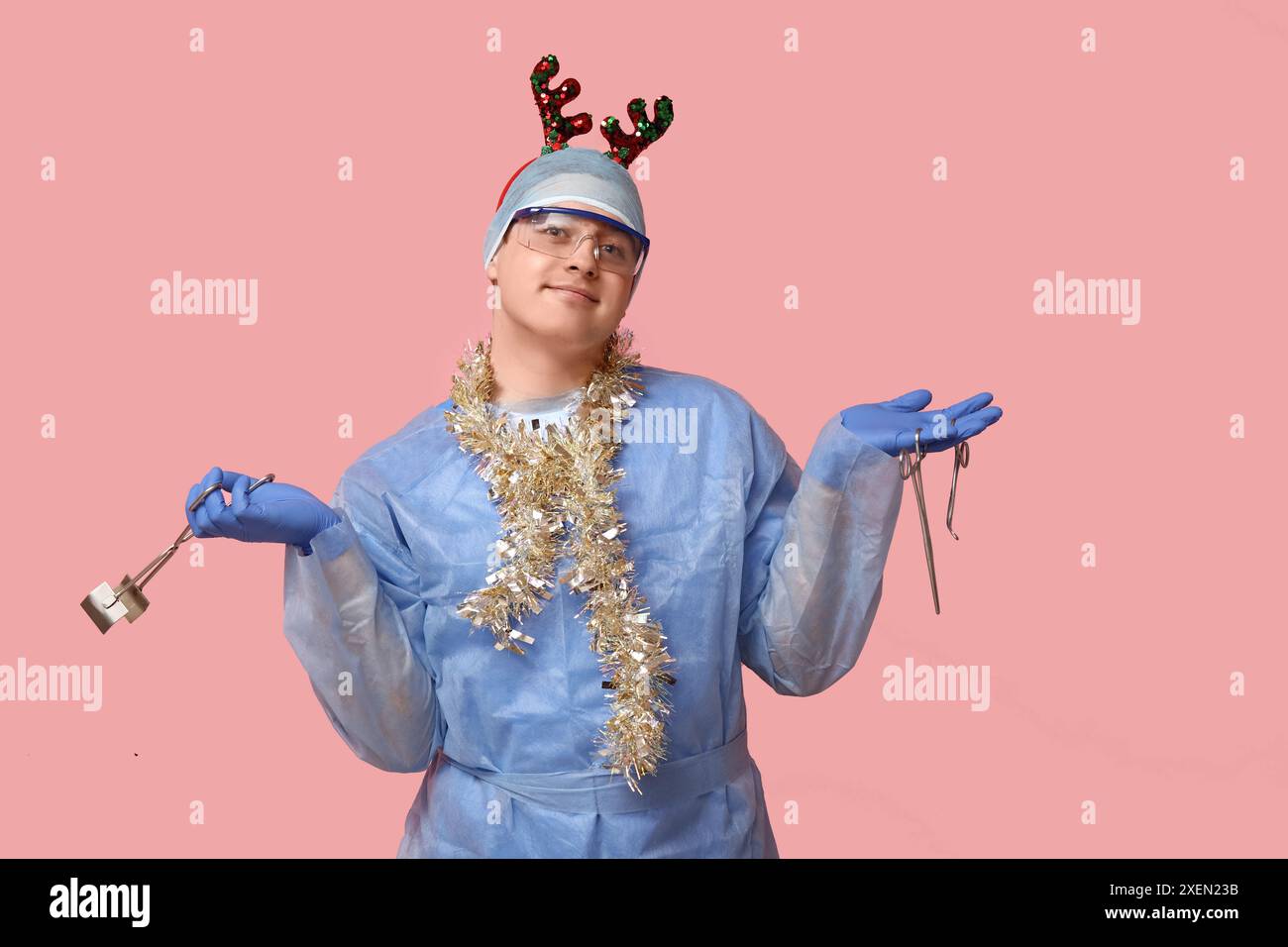 Ritratto di un giovane chirurgo maschio con attrezzi, cerchietto di Natale e cerchietto di renna su sfondo rosa Foto Stock