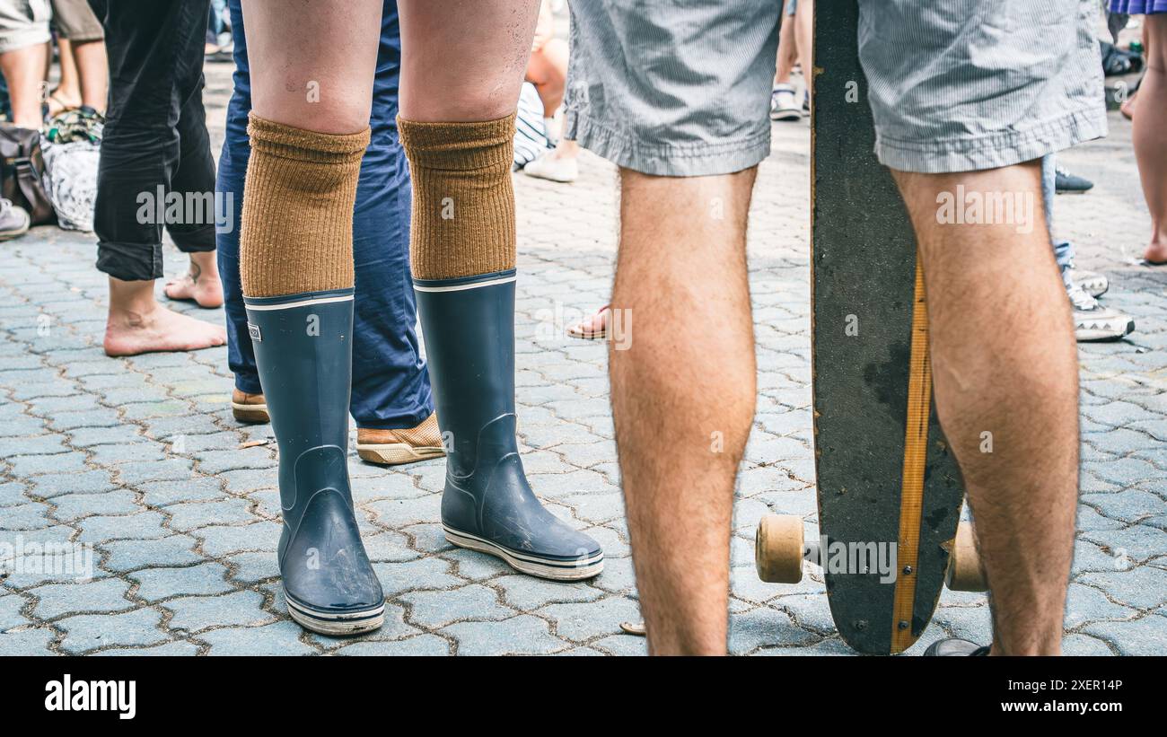 2 paia di gambe l'una di fronte all'altra, una indossa calze al ginocchio con stivali di gomma, l'altra indossa pantaloncini corti, accanto a un longboard Foto Stock