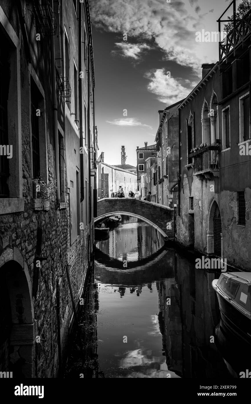 Vista in bianco e nero di uno stretto canale e ponte in pietra a Venezia, Italia, con riflessi sull'acqua e sugli edifici storici che creano un bancomat senza tempo Foto Stock