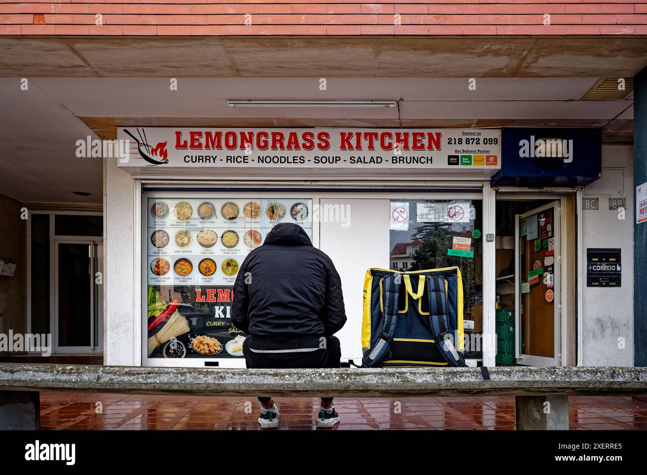 L'addetto alla consegna del cibo siede fuori da un ristorante modesto con il suo zaino isolato al suo fianco, in attesa di un ordine Foto Stock