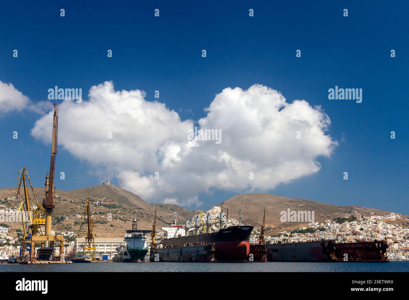 Cantiere navale di Syros con gru torreggianti e navi ancorate sotto un cielo azzurro luminoso con soffici nuvole bianche, che mostrano l'herita industriale e marittima Foto Stock