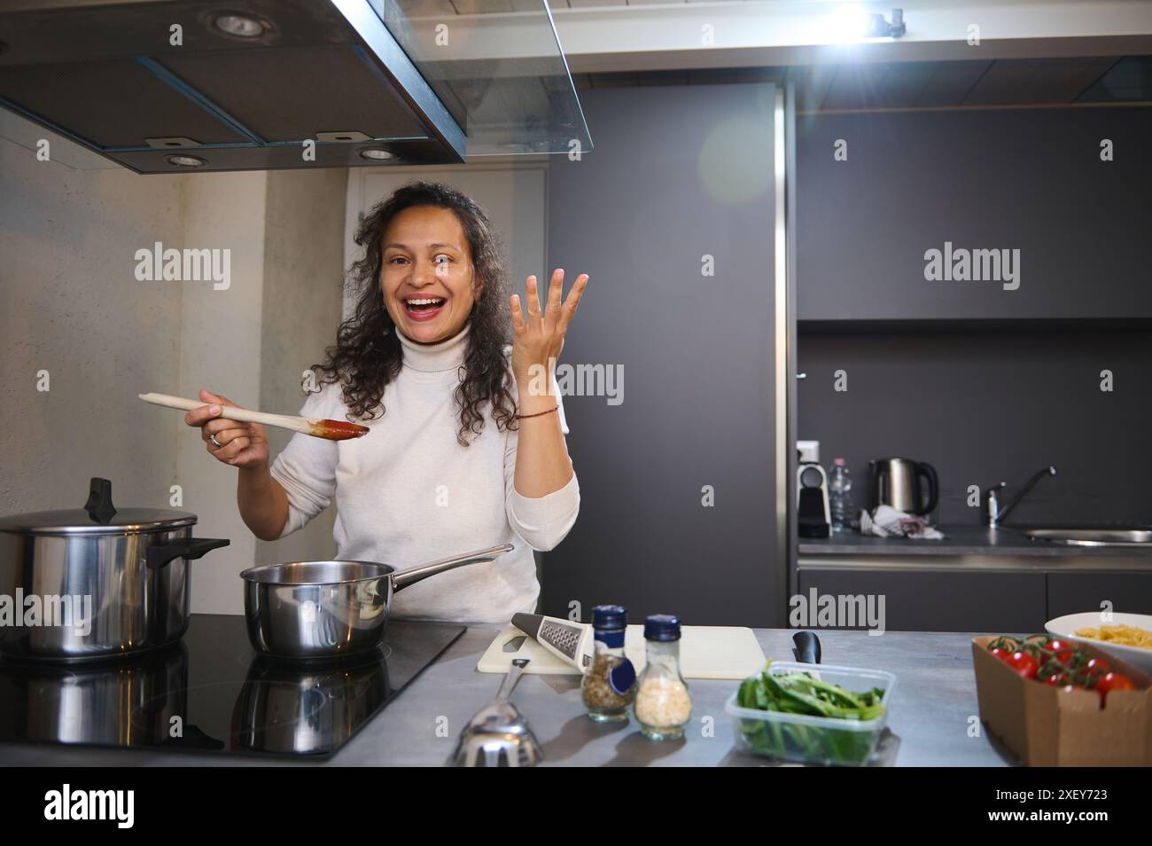 La giovane casalinga, la governante, lo chef, la cuoca, la donna latina che fa il segno della delizia del gusto, assaporando, preparando del cibo delizioso, stando in piedi al bancone della cucina Foto Stock