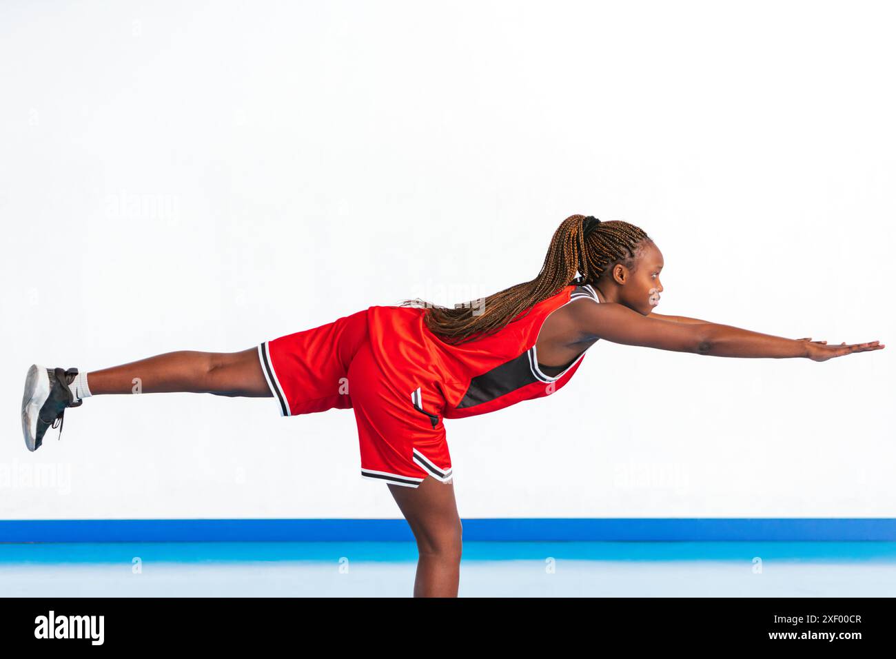 Foto orizzontale una giovane atleta con una divisa sportiva rossa dimostra un esercizio di equilibrio, estendendo una gamba all'indietro e le braccia in avanti per lo stabi Foto Stock