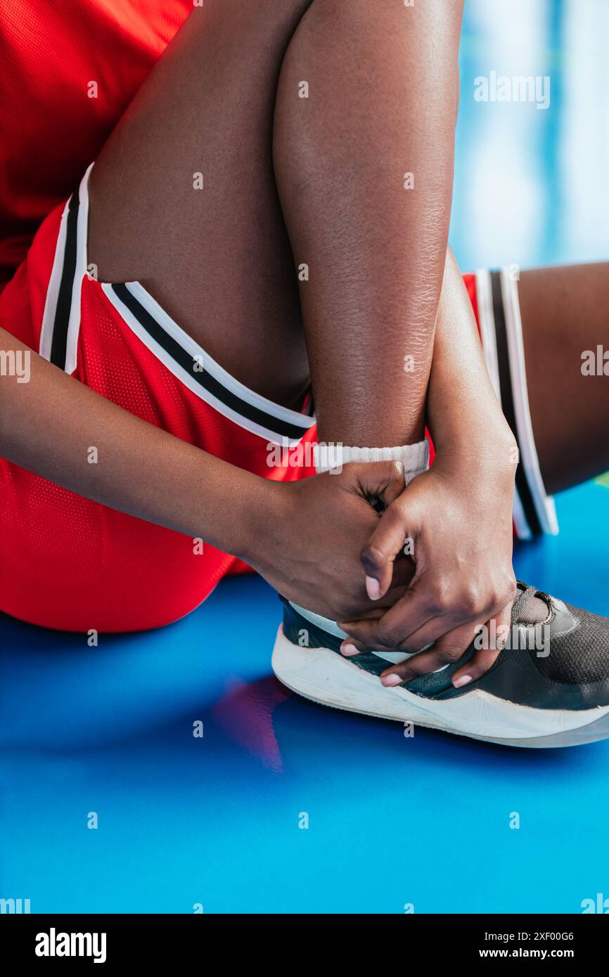 Foto verticale giovane donna in uniforme da basket rossa seduta su un campo blu, tenendo la caviglia ferita con entrambe le mani. L'immagine riflette il dolore e la n Foto Stock