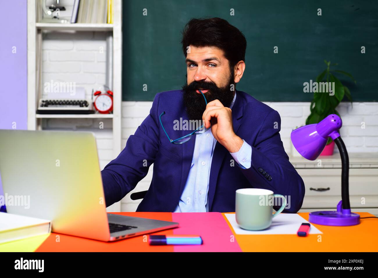 Un insegnante o uno studente sorridente che lavora con un computer portatile in classe. L'insegnante di scuola dà lezione. Uomo d'affari barbato, insegnante online o impiegato siedono Foto Stock