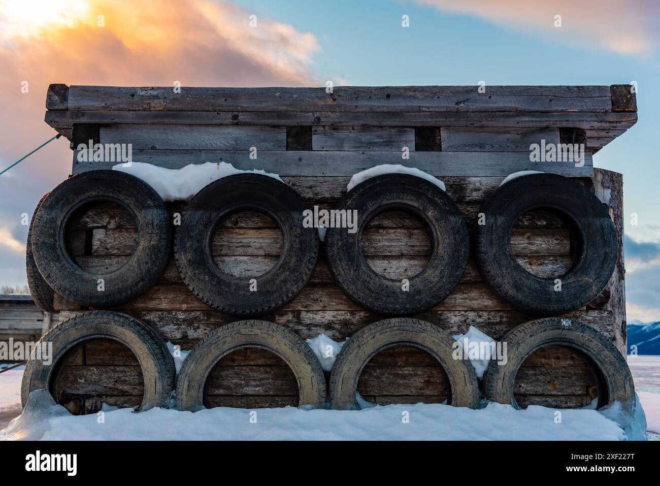 Grandi blocchi di ghiaccio ghiacciati su un porticciolo con pneumatici per auto in inverno. Il tramonto dorato si illumina sul lato di un lago gelido in inverno con il tramonto sullo sfondo. Foto Stock