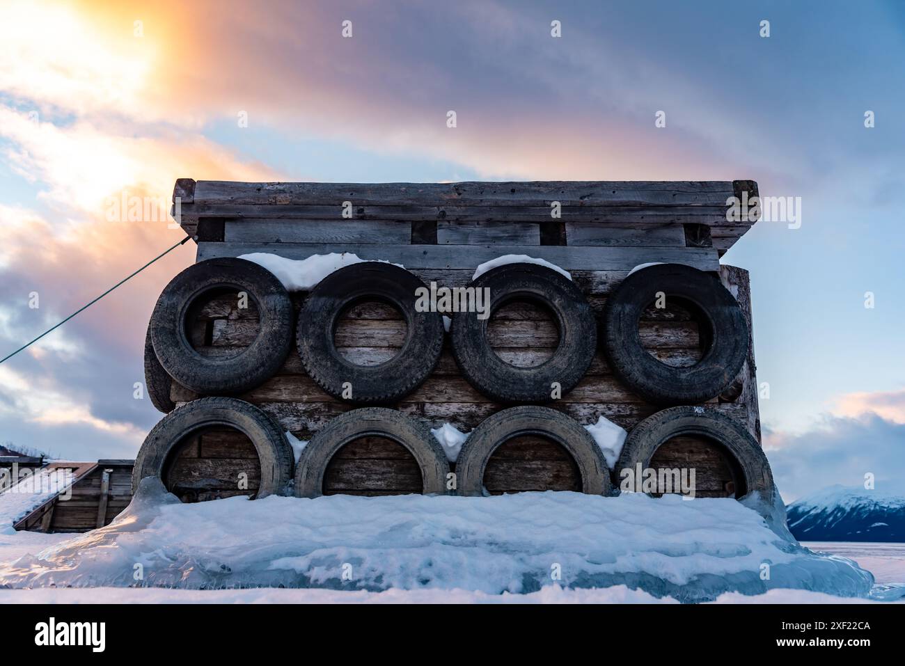 Grandi blocchi di ghiaccio ghiacciati su un porticciolo con pneumatici per auto in inverno. Il tramonto dorato si illumina sul lato di un lago gelido in inverno con il tramonto sullo sfondo. Foto Stock