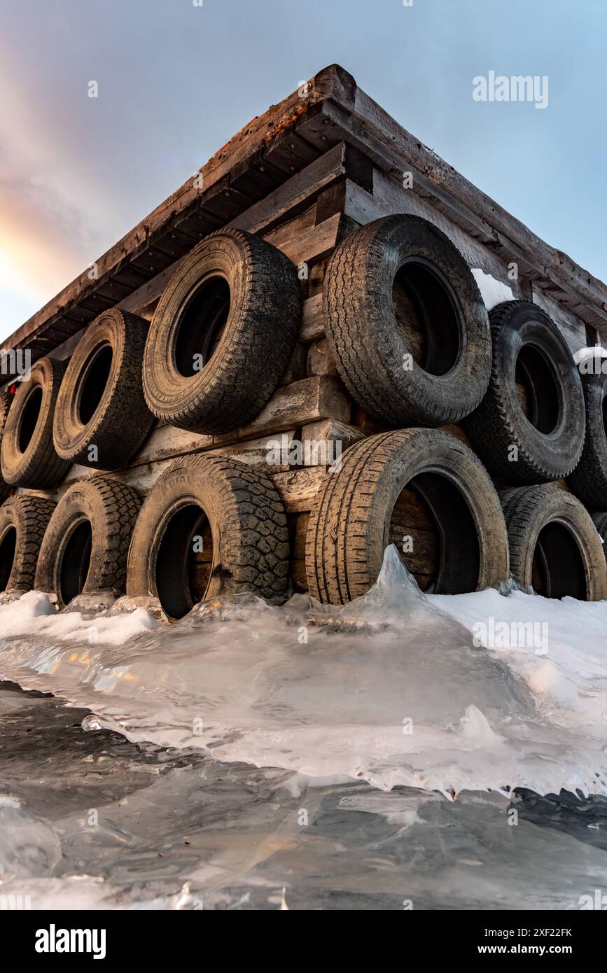 Grandi blocchi di ghiaccio ghiacciati su un porticciolo con pneumatici per auto in inverno. Il tramonto dorato si illumina sul lato di un lago gelido in inverno con il tramonto sullo sfondo. Foto Stock