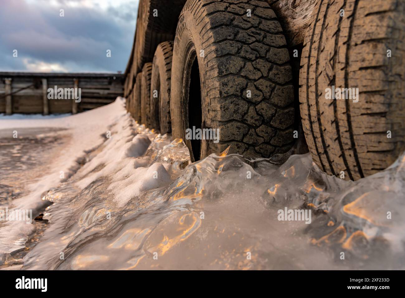 Grandi blocchi di ghiaccio ghiacciati su un porticciolo con pneumatici per auto in inverno. Il tramonto dorato si illumina sul lato di un lago gelido in inverno con il tramonto sullo sfondo. Foto Stock