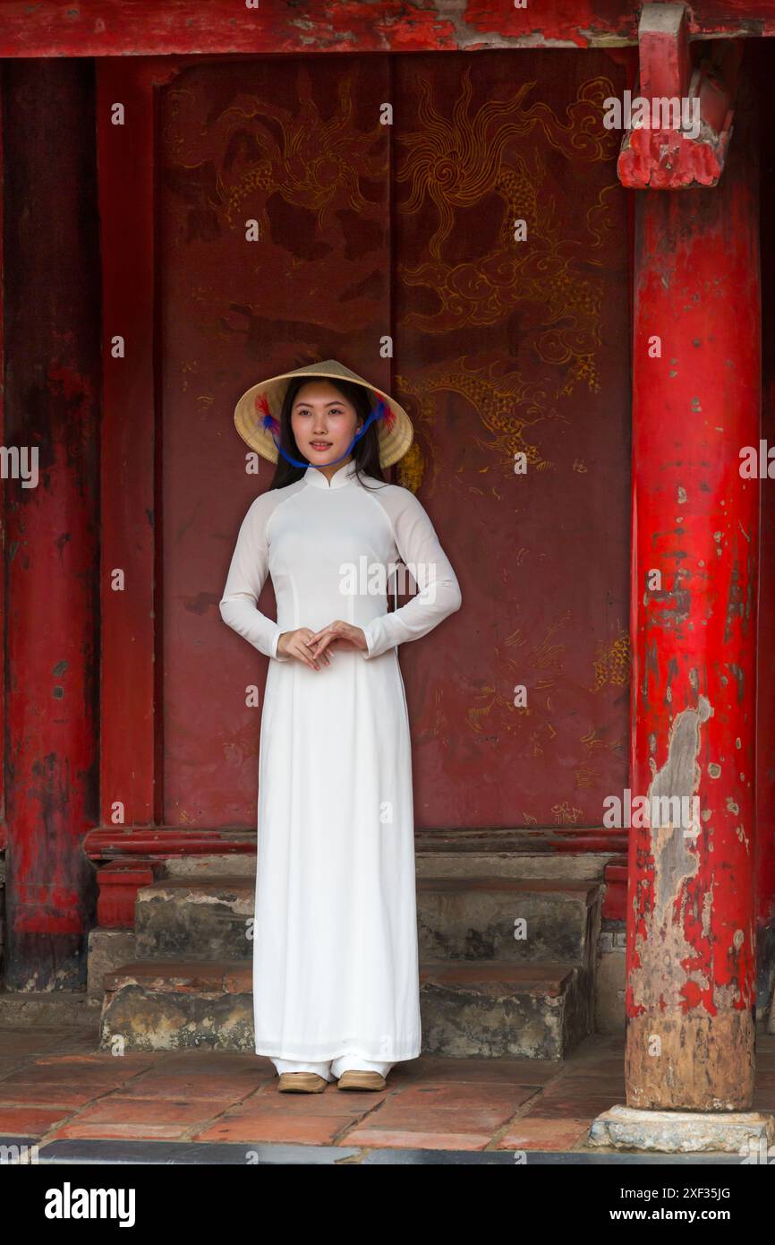 Graziosa giovane donna vietnamita che indossa la tradizionale veste Ao dai e un cappello conico in piedi nella porta ornata del Tempio della Letteratura, Hanoi, Vietnam del Nord Foto Stock