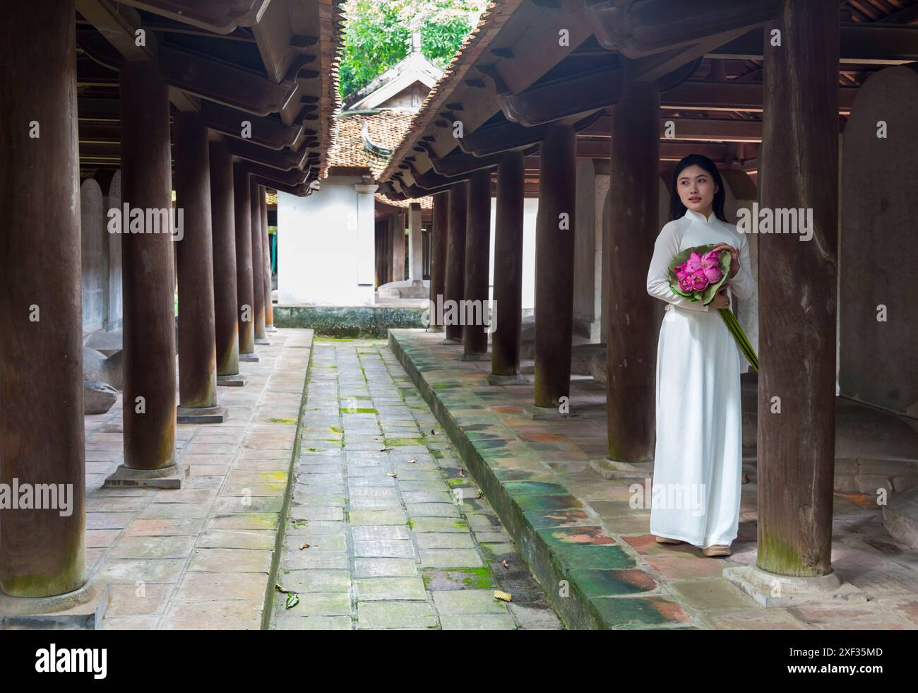 Graziosa giovane donna vietnamita che indossa la tradizionale tunica Ao dai che tiene un mazzo di fiori di loto al Tempio della Letteratura di Hanoi, Vietnam del Nord, Asia Foto Stock