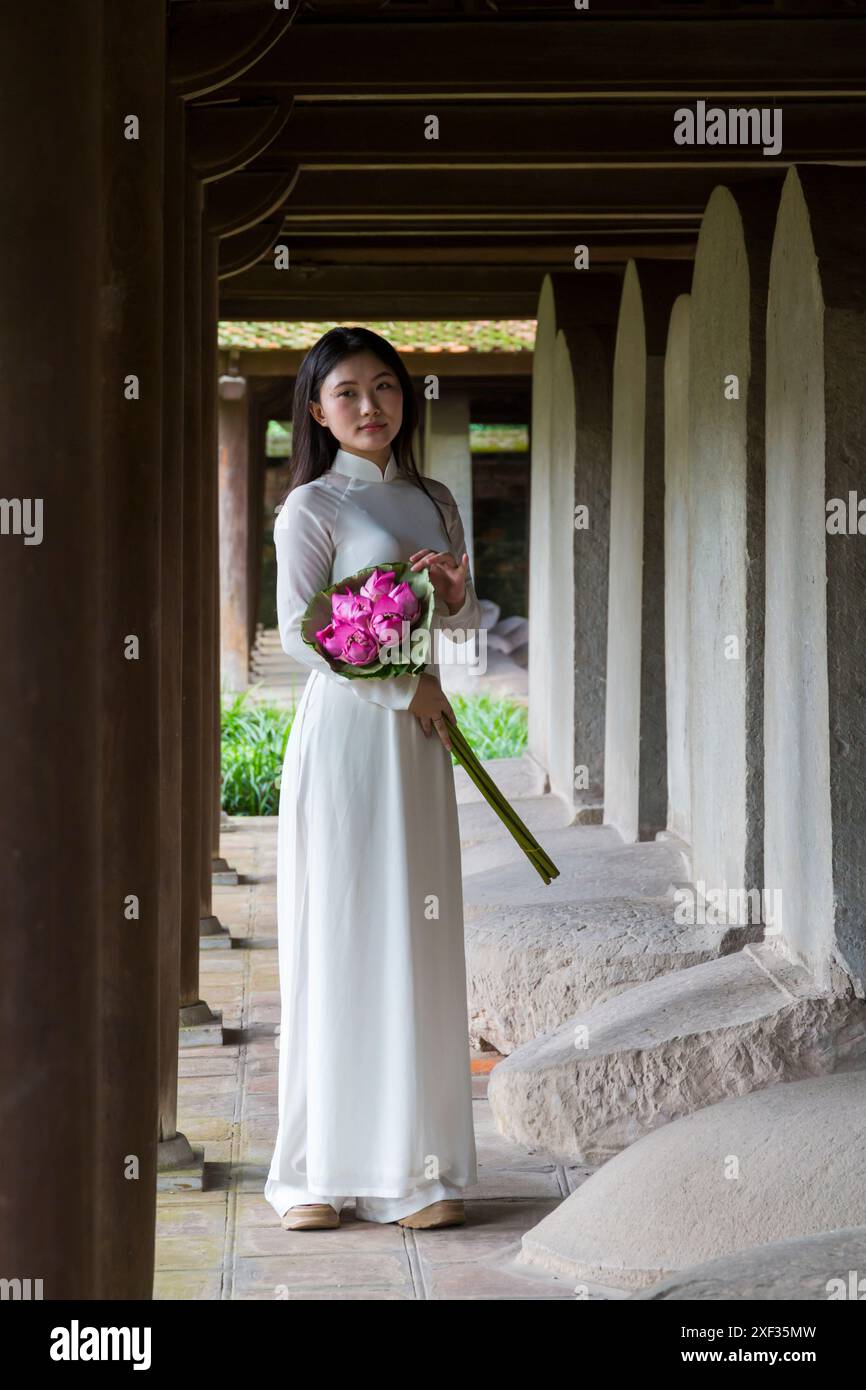 Graziosa giovane donna vietnamita che indossa la tradizionale tunica Ao dai che tiene un mazzo di fiori di loto al Tempio della Letteratura di Hanoi, Vietnam del Nord, Asia Foto Stock