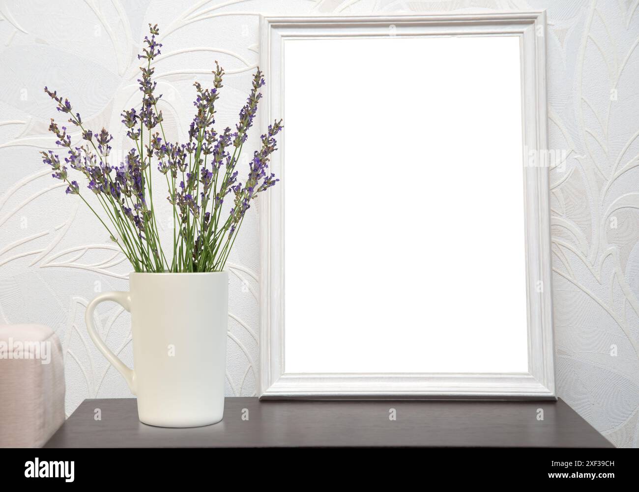 Una tazza bianca con fiori di lavanda si trova su un tavolo accanto a una cornice bianca. Lo sfondo è una parete bianca con un motivo in rilievo. Foto Stock