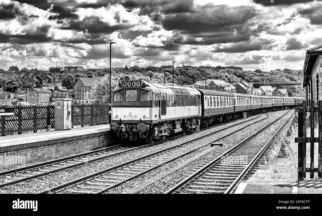 Una vecchia locomotiva diesel della North Yorkshire Moors Railway tira una linea di carrozze mentre si avvicina a una stazione ferroviaria. Un cielo con nuvole è sopra. Foto Stock