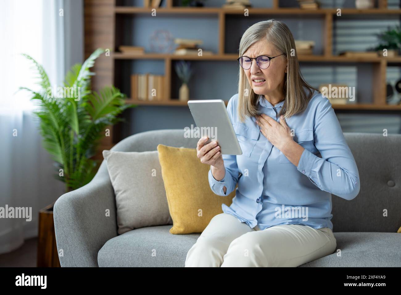 Donna anziana che soffre di dolore toracico durante l'uso della compressa per consultare il medico online. Si siede su un divano in un elegante soggiorno, sembra preoccupata e tiene il petto. Foto Stock