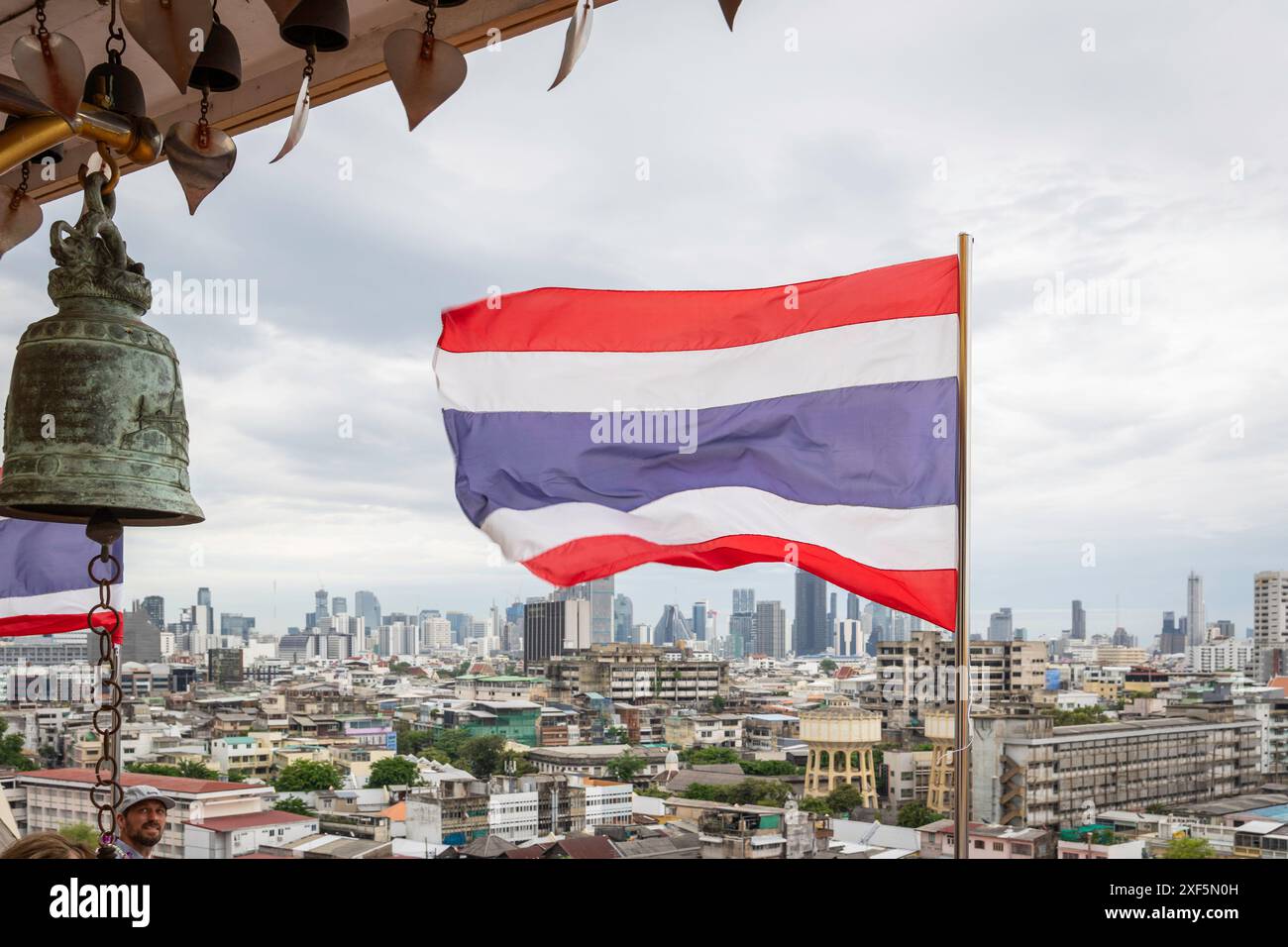 Bangkok, Thailandia. 25 giugno 2024. La bandiera nazionale thailandese si vede sventolare vicino a una campana buddista, con una vista aerea di Bangkok sullo sfondo, in cima al Monte d'Oro, al Tempio Wat Saket. Bangkok è un crogiolo di culture, religioni e costumi. Prevalentemente buddista, ha una storia di accoglienza dei coloni con credenze diverse. Bangkok ospita santuari, templi e chiese dove persone di varie fedi pregano per fortuna e fortuna. Credito: SOPA Images Limited/Alamy Live News Foto Stock