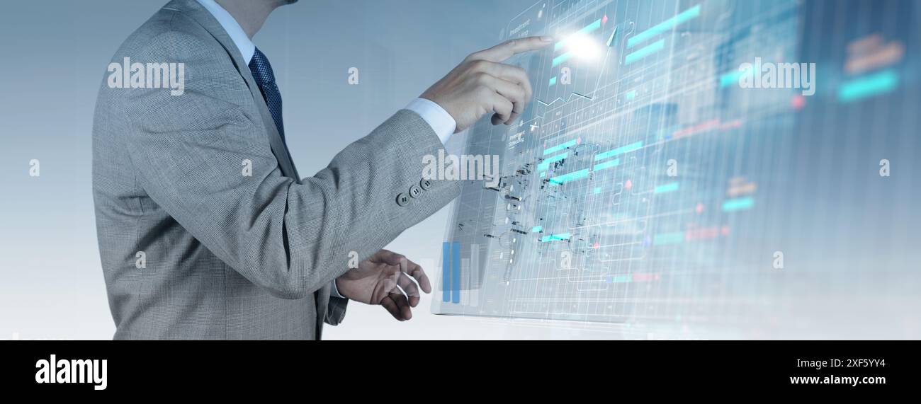 Un uomo d'affari in una tuta grigia interagisce con un'interfaccia digitale futuristica che visualizza dati, grafici e pianificazioni complessi. Questa immagine evidenzia la u Foto Stock