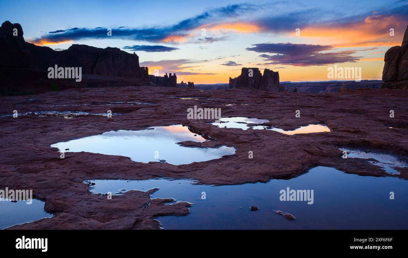La notte scende al Parco Nazionale degli Arches con le pozzanghere di acqua piovana nelle rocce al crepuscolo, mentre le nuvole si illuminano di colori caldi Foto Stock