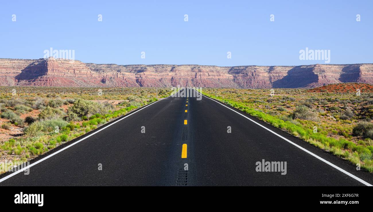 Panorama dell'autostrada desertica con asfalto nero fresco e rigoglioso bordo verde che conduce alle scogliere di arenaria rossa nello Utah meridionale Foto Stock