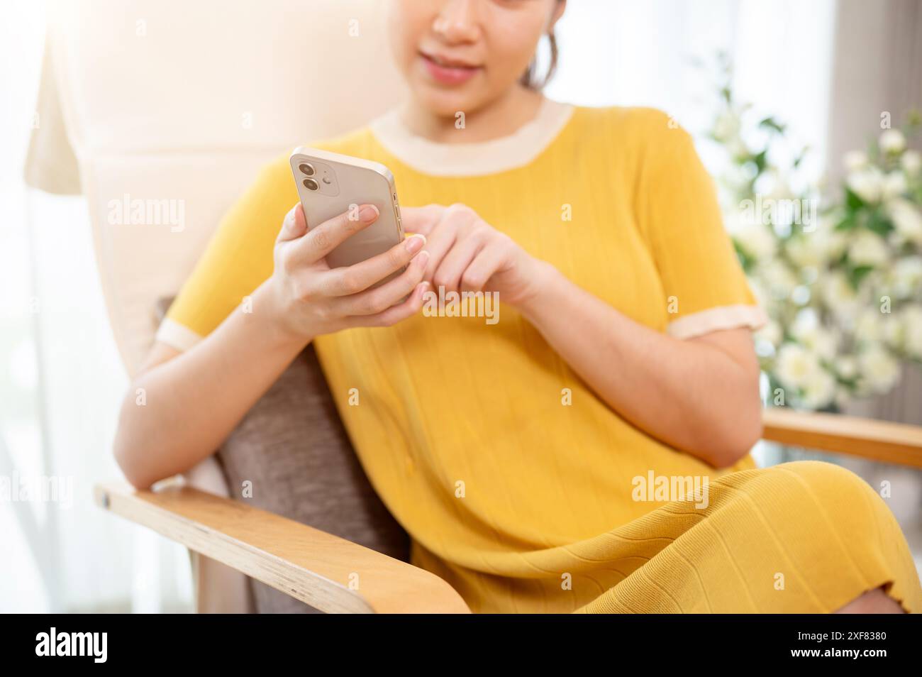 Un'immagine ritagliata di una donna asiatica in un abito giallo sta usando il suo smartphone mentre si rilassa su una poltrona nel soggiorno. scorrere sul telefono, leggere Foto Stock