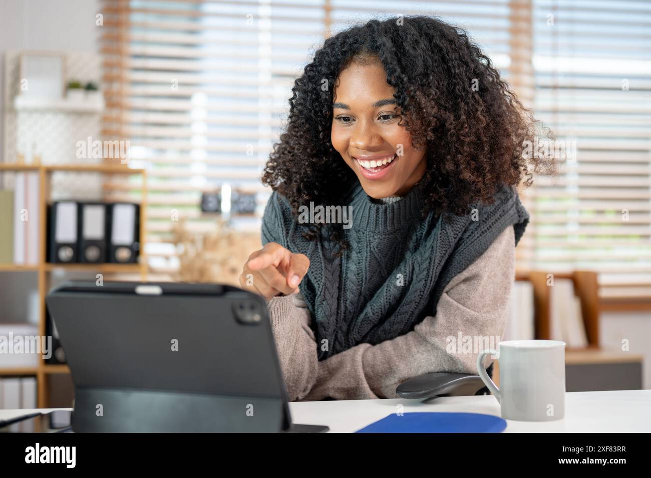 Un'attraente e allegra donna nera in un comodo maglione a maglia punta il dito sullo schermo di un tablet mentre si tiene una riunione online con il suo collea Foto Stock